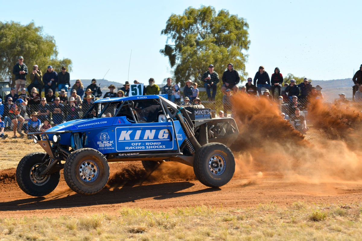 Blue off-road vehicle racing on a dirt track with spectators watching.